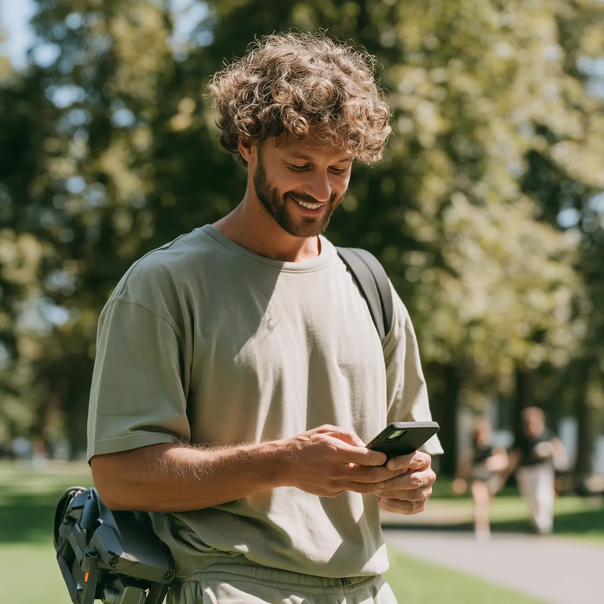 A smiling person in a park looks at their phone, with a drone bag at their side.